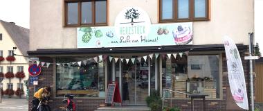 Außenansicht des Herzstück Diedorf mit großer Glasfront, darüber ein Schild mit dem Text "HERZSTÜCK ... aus Liebe zur Heimat!" und dem charakteristischen Baum-Logo, vor dem Laden steht eine Person mit Lastenrad